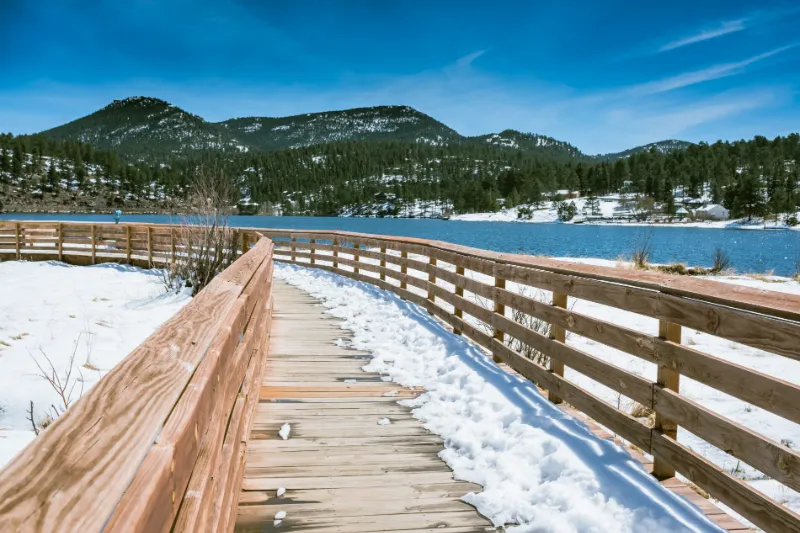 VIEW WOODEN WALK WAY WITH BACKGROUND OF BRIGHT BLUE SKY AND SNOW ON GROUND OF EVERGREEN LAKE IN LATE WINTER / COLORADO / DENVER